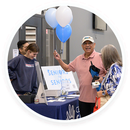 A man with blue and white balloons talks at a seniors' info table for youth volunteer programs Texas.